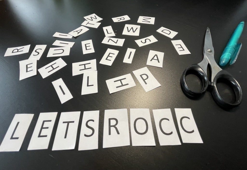 Hand-cut paper letter tiles spelling out LETSROCC on a dark table, with scattered spare letters and scissors. The moment the company name was born.